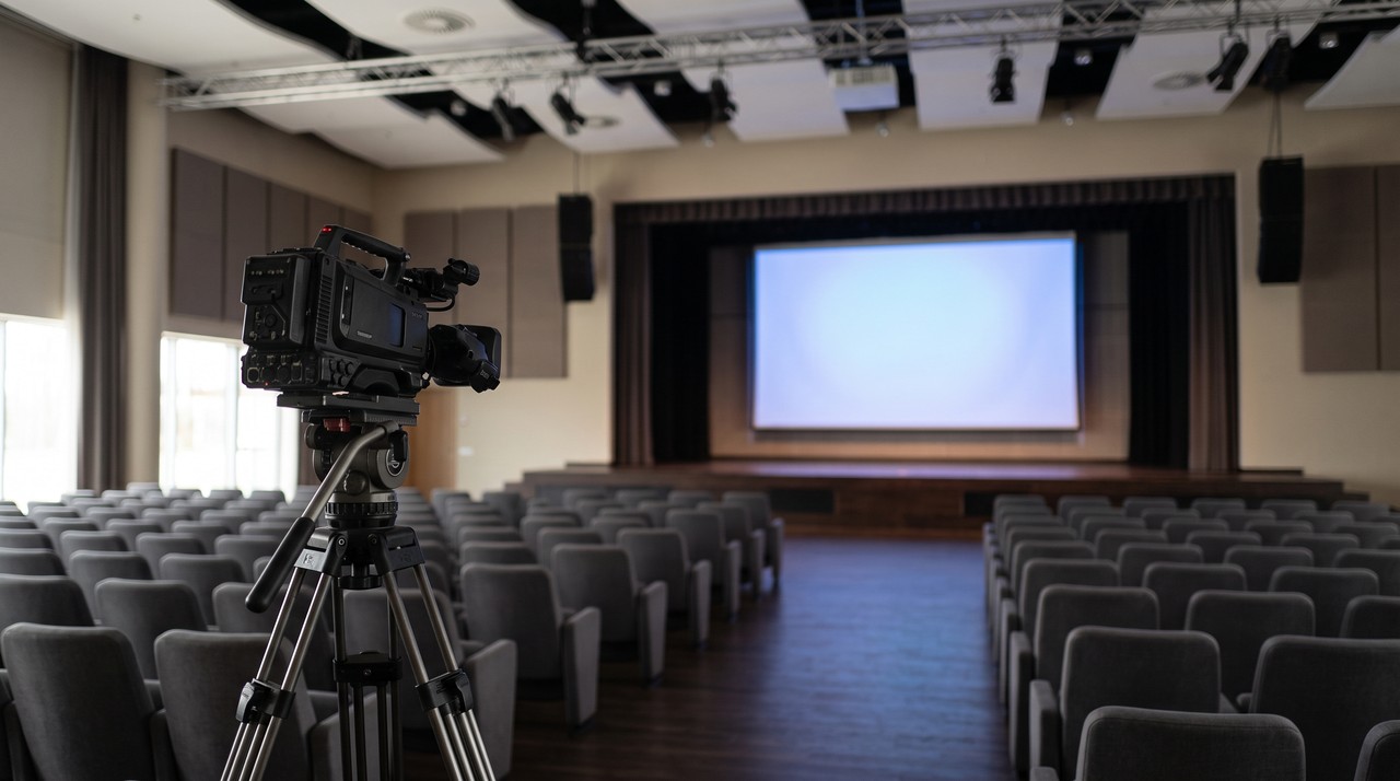 A video camera on a tripod facing a conference stage and its projection screen